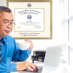 Man working at desk on computer with coffee cup in one hand and a National Board of Certification For Medical Interpreters Certificate Frame on the wall behind him.