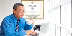 Man working at desk on computer with coffee cup in one hand and a National Board of Certification For Medical Interpreters Certificate Frame on the wall behind him.