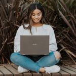 Woman sitting cross-legged in jeans and sweater on a wooden walkway working on her laptop with ornamental grasses behind her.