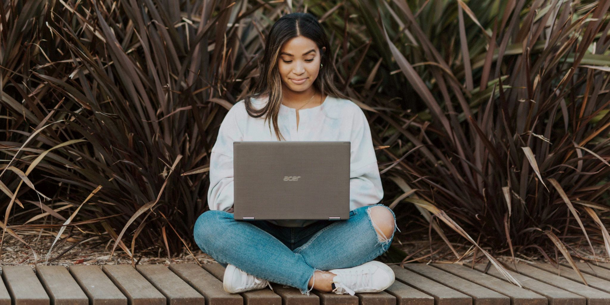 Woman sitting cross-legged in jeans and sweater on a wooden walkway working on her laptop with ornamental grasses behind her.