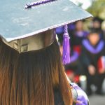 Closeup of the back of a grad wearing a mortarboard with tassel and grad gown facing crowd of graduates.