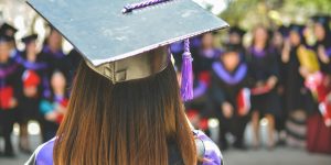 Closeup of the back of a grad wearing a mortarboard with tassel and grad gown facing crowd of graduates.