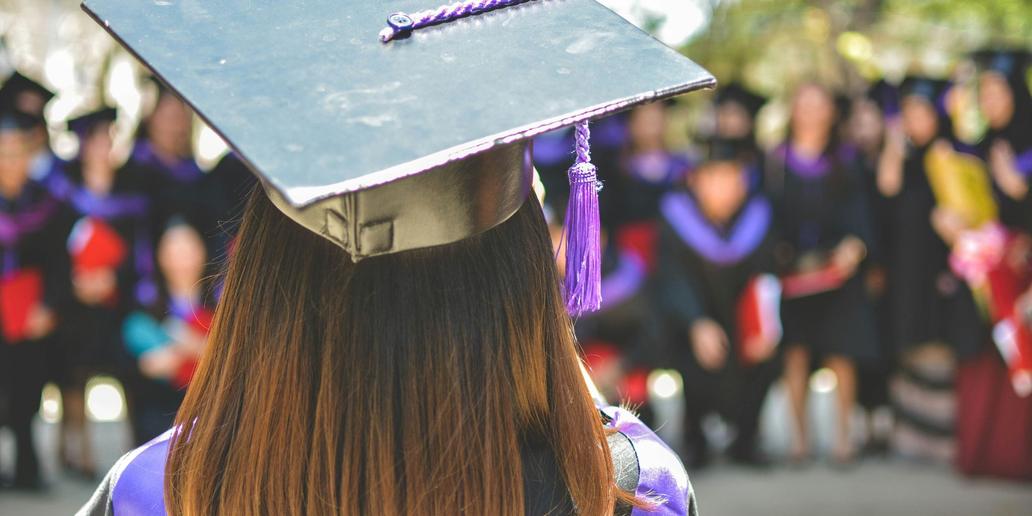 Closeup of the back of a grad wearing a mortarboard with tassel and grad gown facing crowd of graduates.