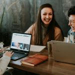 Three college friends laughing while working on laptops at cafe.
