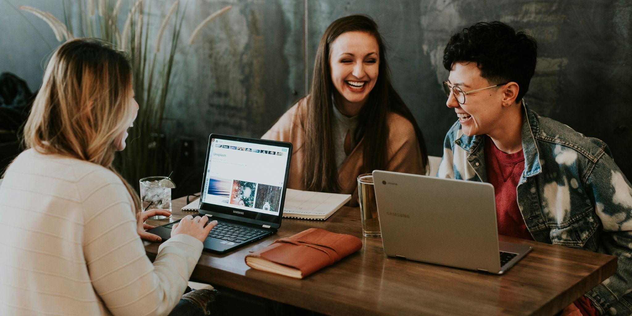 Three college friends laughing while working on laptops at cafe.