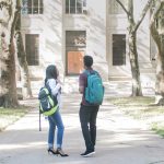 Two students outside on college campus in front of building chatting.