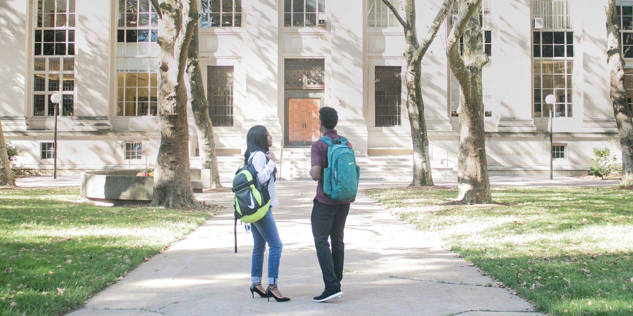 Two students outside on college campus in front of building chatting.
