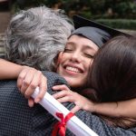 Smiling graduate holding her diploma and wearing her grad cap hugging her parents on campus.