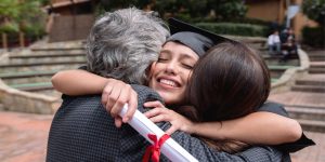 Smiling graduate holding her diploma and wearing her grad cap hugging her parents on campus.