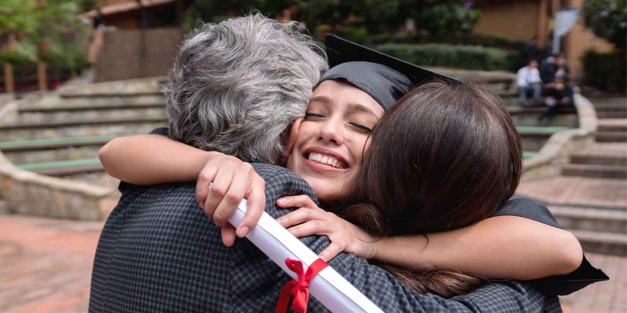 Smiling graduate holding her diploma and wearing her grad cap hugging her parents on campus.