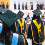 The backs of master's graduates wearing doctoral sashes and hoods as they proceed toward the graduation stage.
