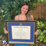 Smiling woman holding University of Florida diploma frame in front of garden and fence in backyard.