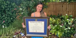 Smiling woman holding University of Florida diploma frame in front of garden and fence in backyard.
