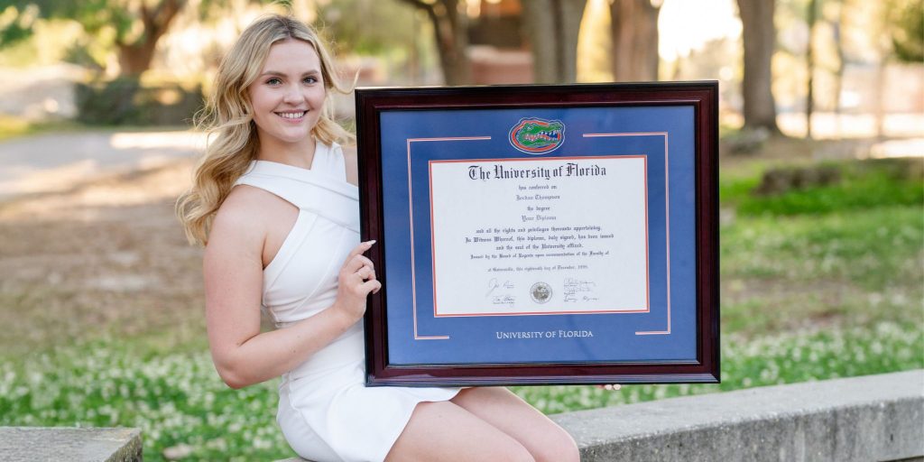 Woman in white dress sitting on stone wall holding a blue University of Florida diploma frame with a Gator logo medallion.