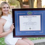 Woman in white dress sitting on stone wall holding a blue University of Florida diploma frame with a Gator logo medallion.