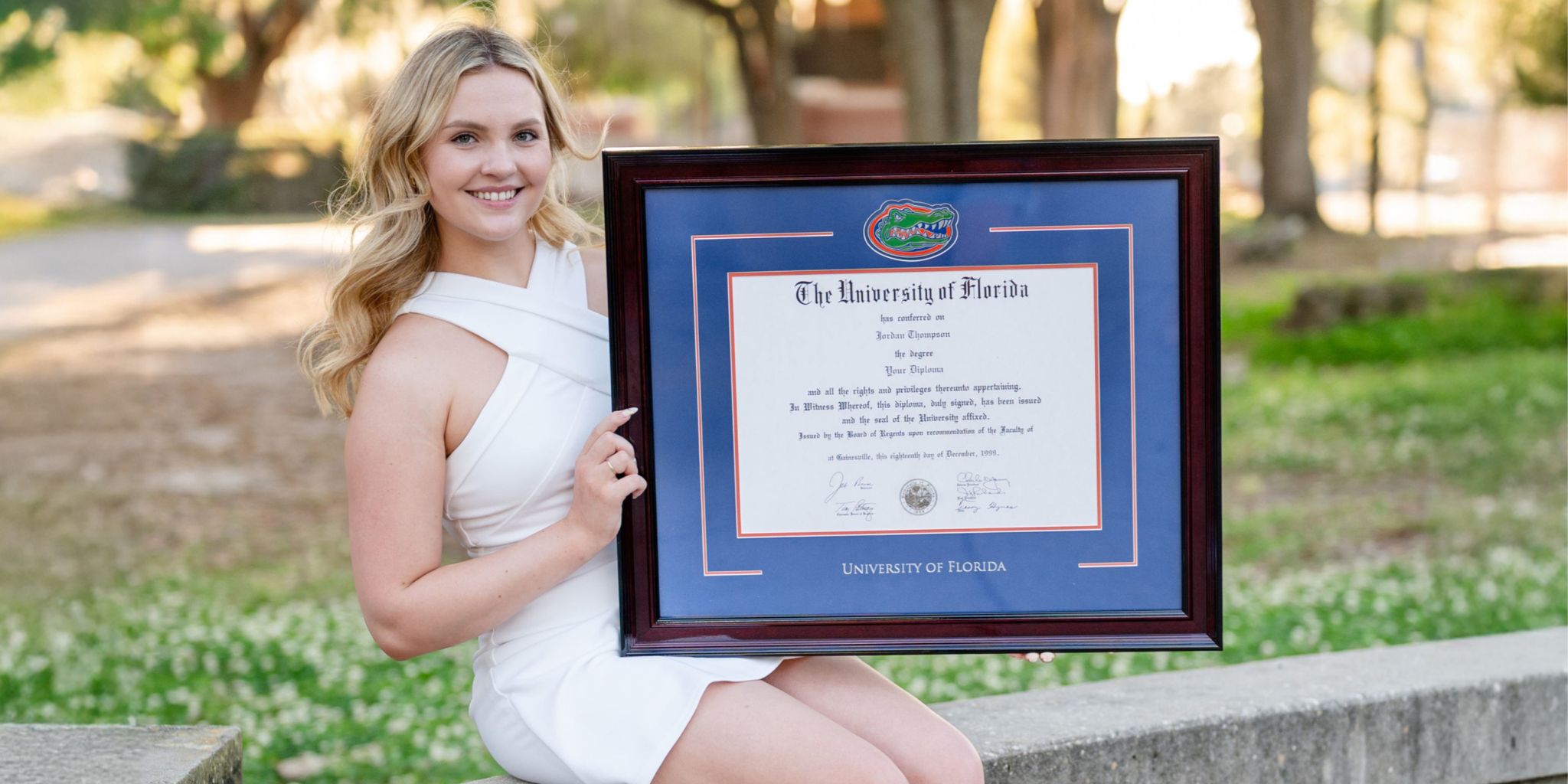Woman in white dress sitting on stone wall holding a blue University of Florida diploma frame with a Gator logo medallion.