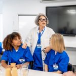 Medical professor wearing lab coat and stethoscope in classroom speaking to medical students in blue scrubs.