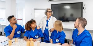 Medical professor wearing lab coat and stethoscope in classroom speaking to medical students in blue scrubs.