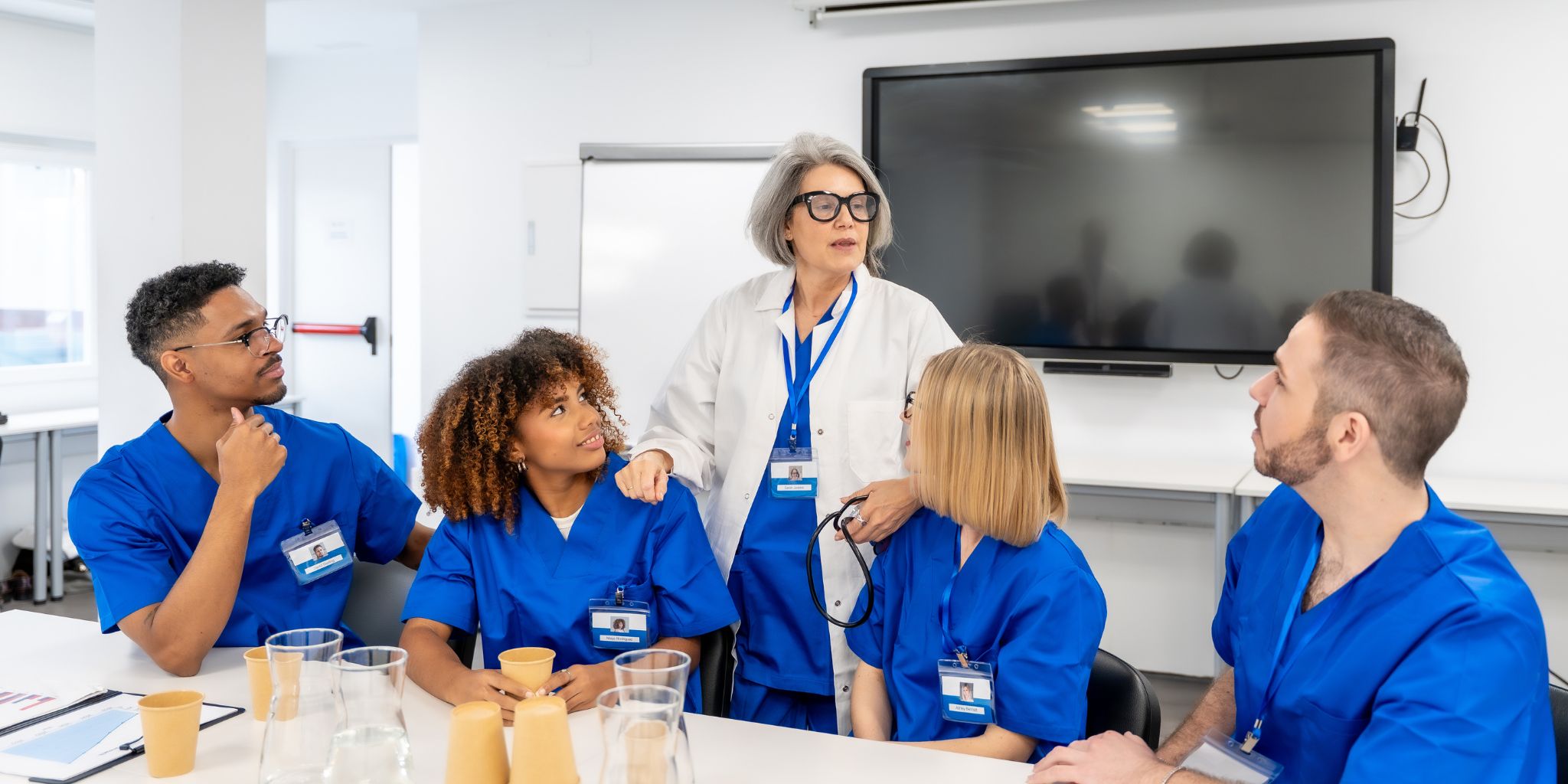 Medical professor wearing lab coat and stethoscope in classroom speaking to medical students in blue scrubs.