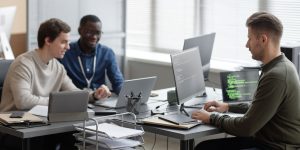 Three IT workers working on their laptops in an office at a communal table.