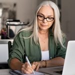 Woman with glasses and gray hair holding pen and working in notebook in front of laptop in a work office.
