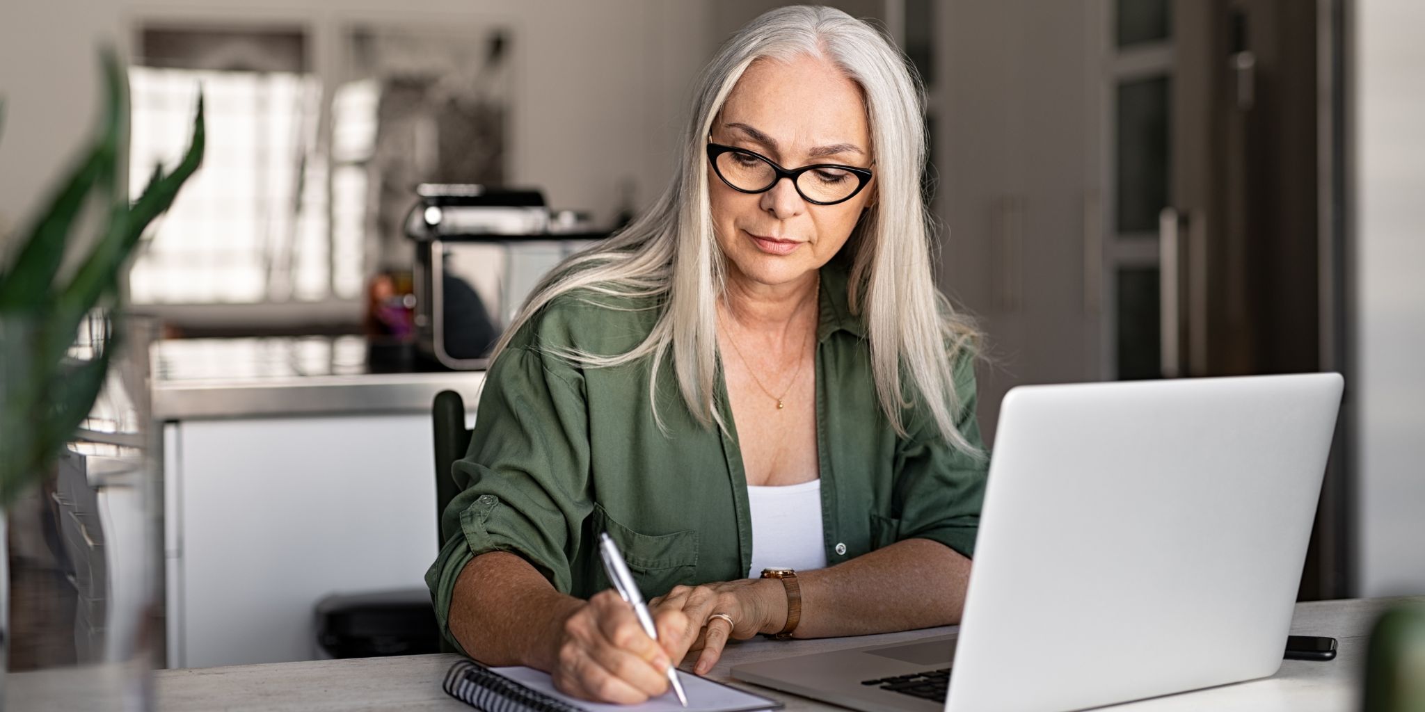 Woman with glasses and gray hair holding pen and working in notebook in front of laptop in a work office.