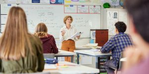 Teacher in front of class lecturing while holding a clipboard.