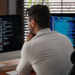 Back of a person at a desk in an office with blinds working on two laptops.
