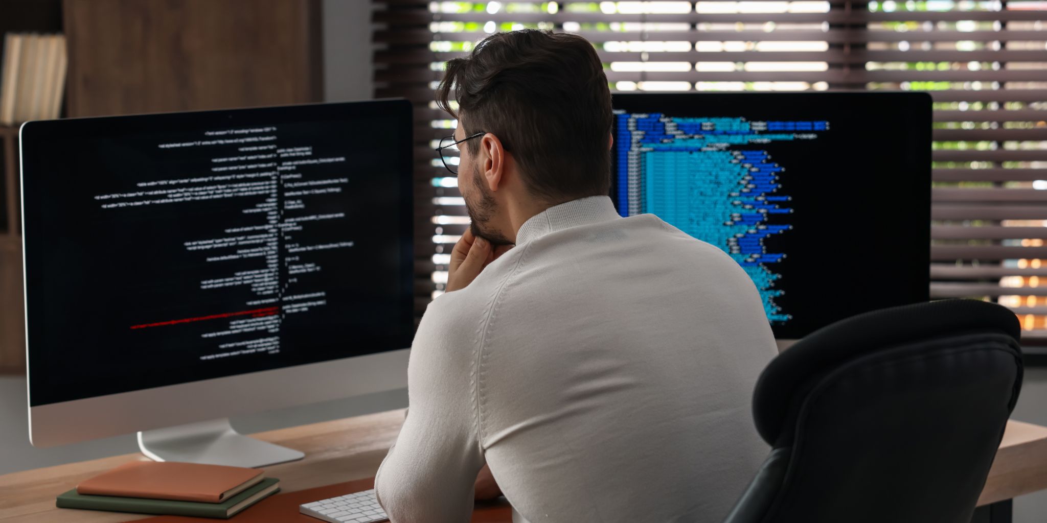 Back of a person at a desk in an office with blinds working on two laptops.