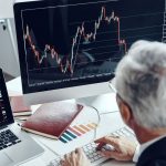 Back of a person in a suit with gray hair working at a desk with two computers with graphs on screen.