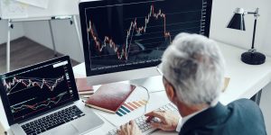 Back of a person in a suit with gray hair working at a desk with two computers with graphs on screen.