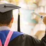 Back of graduate in cap, gown, and sash holding a rolled diploma outdoors.