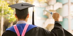 Back of graduate in cap, gown, and sash holding a rolled diploma outdoors.