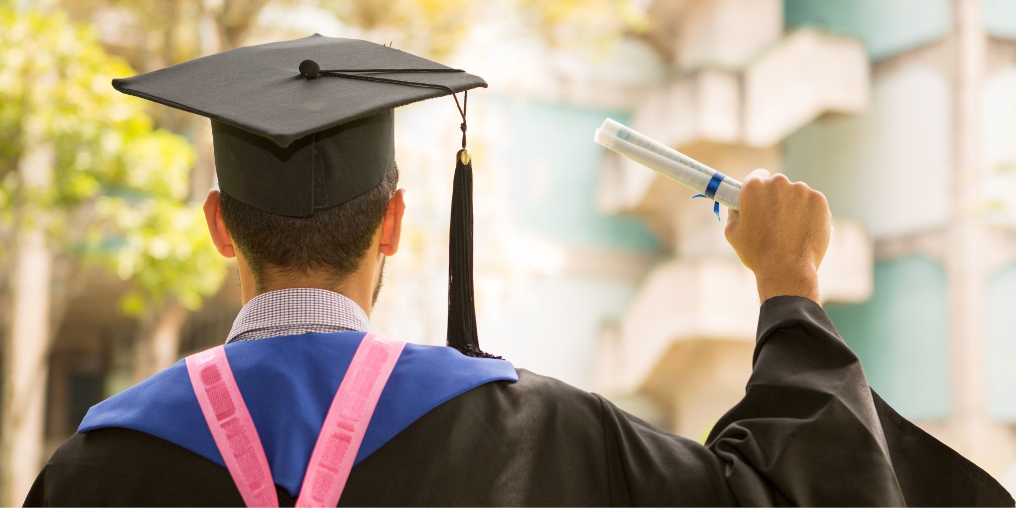 Back of graduate in cap, gown, and sash holding a rolled diploma outdoors.