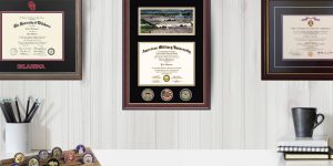 Home office area with Oklahoma State University diploma frame, American Military University diploma frame with challenge coins, and Purple Heart frame over desk with coffee cup and pencil holder.