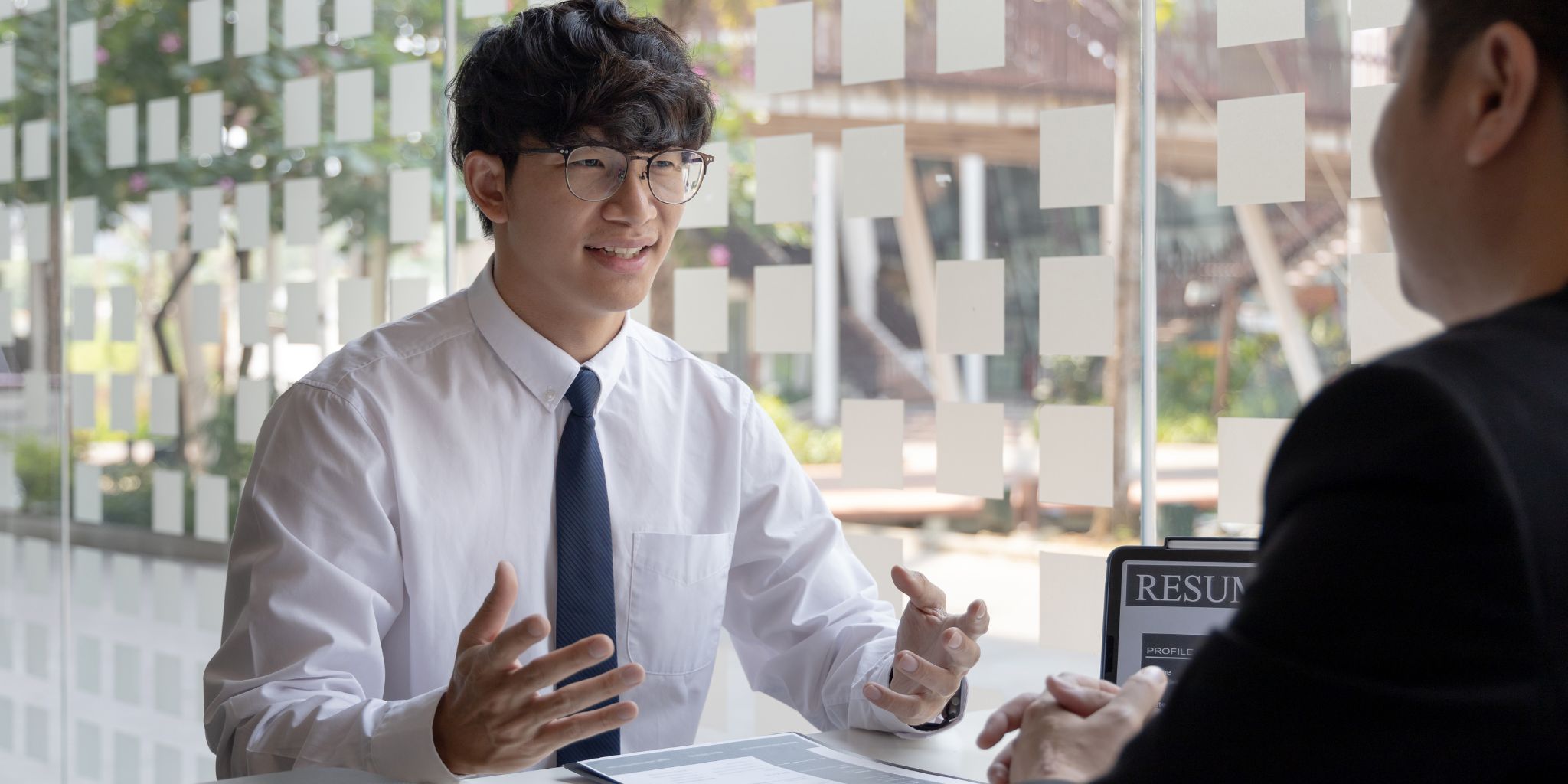 Person in tie and glasses getting interviewed for a job near an office window with resume on laptop screen.