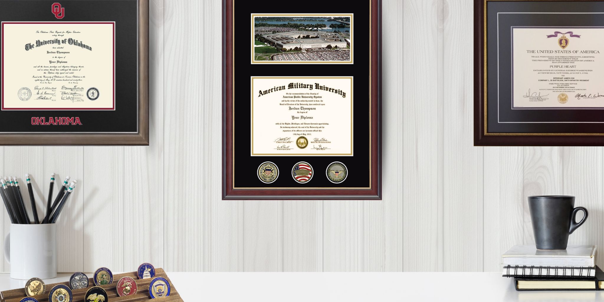 Home office area with Oklahoma State University diploma frame, American Military University diploma frame with challenge coins, and Purple Heart frame over desk with coffee cup and pencil holder.