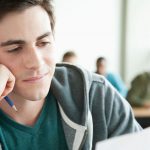 College student sitting in classroom looking at sheet of paper, holding pencil in hand, looking pensive.