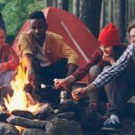 College kids outdoors around campfire roasting marshmallows on sticks with red tent behind them.