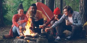 College kids outdoors around campfire roasting marshmallows on sticks with red tent behind them.