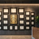 Waiting room with upscale gray wall featuring a collection of framed credentials and business awards with person answering phone at desk nearby.