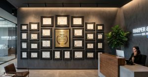 Waiting room with upscale gray wall featuring a collection of framed credentials and business awards with person answering phone at desk nearby.