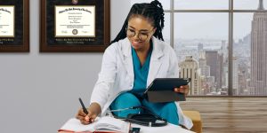 Nurse working at desk writing notes and wearing aqua scrubs in office with a Rutgers School of Nursing degree frame on wall behind her.
