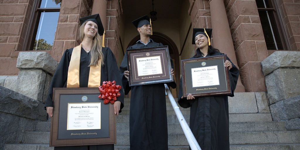 Three graduates in regalia on steps of college holding college diploma frames.