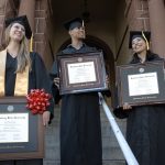 Three graduates in regalia on steps of college holding college diploma frames.