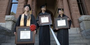 Three graduates in regalia on steps of college holding college diploma frames.