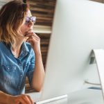 Woman working on computer at desk wearing glasses and a denim blue shirt.