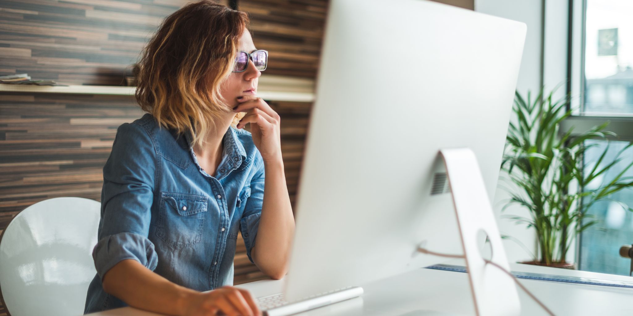 Woman working on computer at desk wearing glasses and a denim blue shirt.