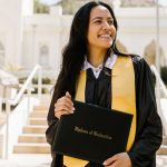 Graduate on campus wearing a yellow graduation stole and holding a diploma cover that says "Diploma of Graduation".
