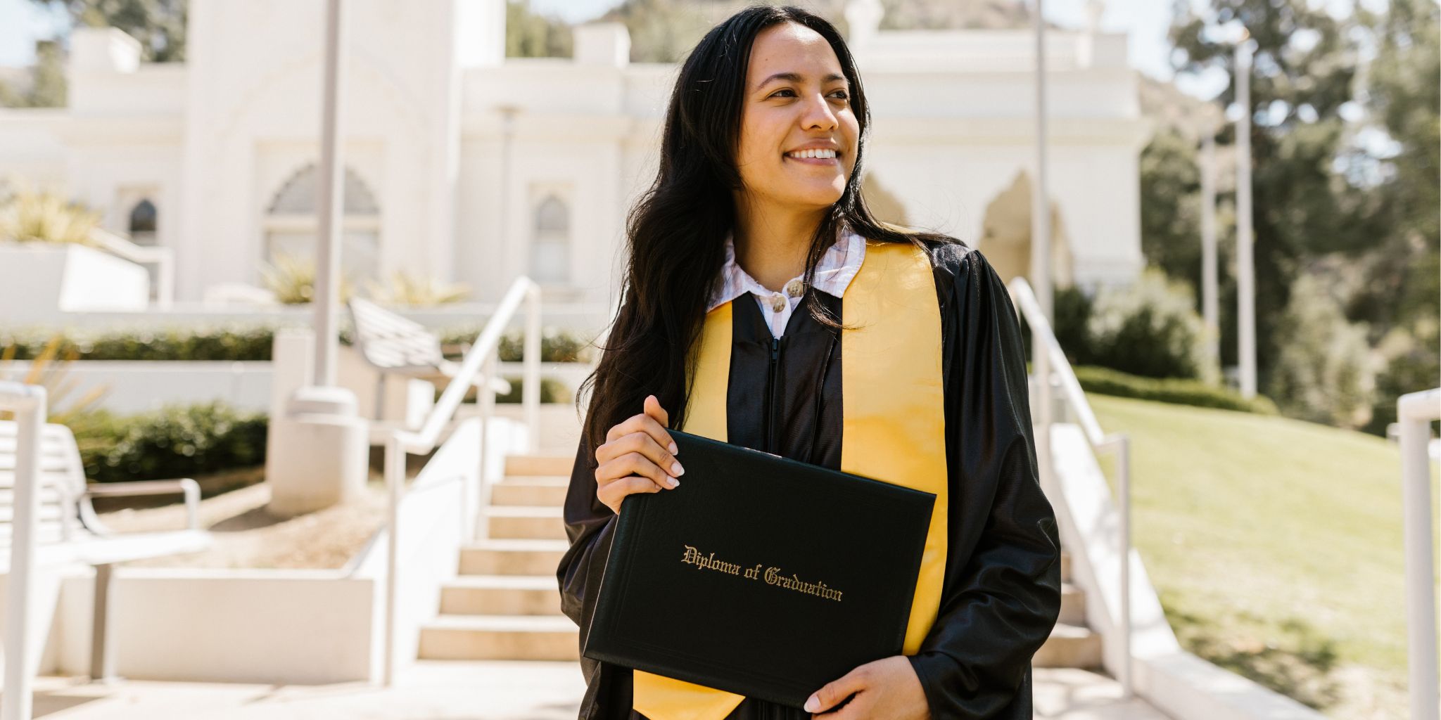 Graduate on campus wearing a yellow graduation stole and holding a diploma cover that says "Diploma of Graduation".